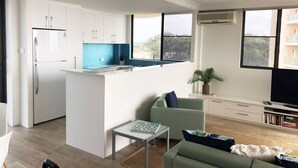 A modern apartment with a kitchen, dining area, and living room. It features a large window, a flat-screen TV, and a bookshelf. - Pinnacle Unit 701 (Forster)