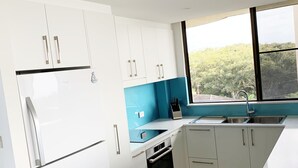 A modern kitchen with white cabinets, a stainless steel refrigerator, a blue backsplash, and a large window offering a view of greenery outside. - Pinnacle Unit 701 (Forster)