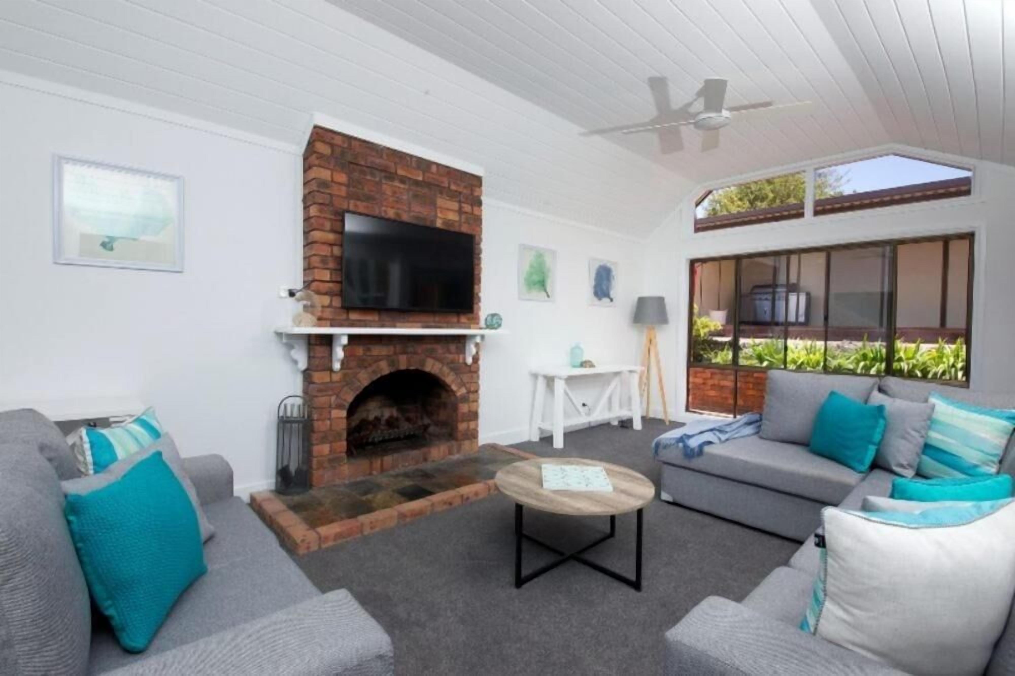 A living room with a brick fireplace, grey sofas with teal cushions, a round coffee table, and a ceiling fan.