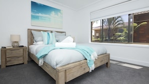 Quarto com cama de madeira, mesa de cabeceira, luminária, janela com vista para palmeiras e um quadro de uma praia.