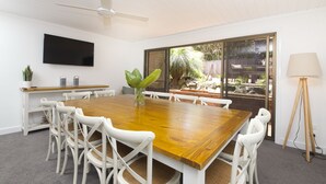 Um quarto espaçoso com uma grande mesa de madeira, cadeiras brancas, um ventilador de teto, televisão e vista para um jardim exterior através de uma porta de correr de vidro.
