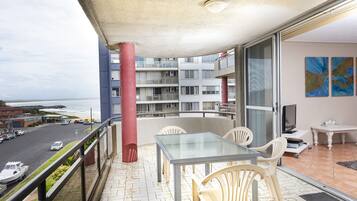 A balcony with a glass table and chairs, overlooking a beach and a multi-story building.