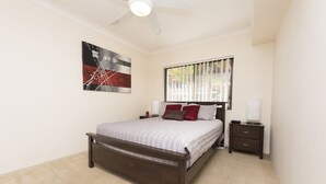 Ein Schlafzimmer mit einem Bett, einem Nachttisch, einem Deckenventilator, einem Fenster mit Jalousien und einem Wandbild.