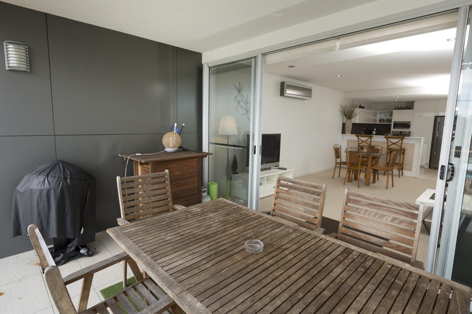 Zona pranzo con tavolo e sedie in legno, barbecue coperto, e vista su una cucina e un soggiorno moderni.