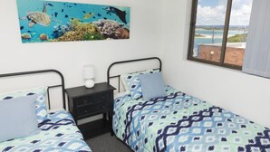 A bedroom with two beds, a nightstand, a lamp, and a large window. - Wharf Street Vista (Forster)