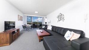 A modern living room with a black leather sofa, wooden coffee table, and a TV on a wooden cabinet. There is a dining area with a table and chairs, and a large window offering a view of the outdoors. - Wharf Street Vista (Forster)