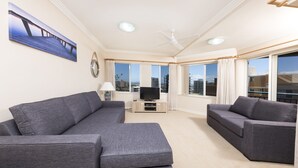 A modern living room with two grey sofas, a flat-screen TV on a wooden stand, and large windows offering a city view. - Whitesands Unit 602 (Forster)