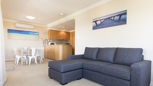 A living room with a grey sectional sofa, a dining area with white chairs, a kitchen with wooden cabinets, and a beach-themed painting on the wall. - Whitesands Unit 602 (Forster)