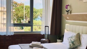 A hotel room with a bed, a window with curtains, a suitcase, and a plant. - Atico Hotel (Desenzano del Garda)