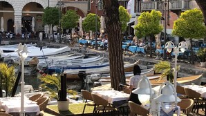A riverside dining area with tables and chairs, boats docked along the water, and buildings in the background. - Atico Hotel (Desenzano del Garda)