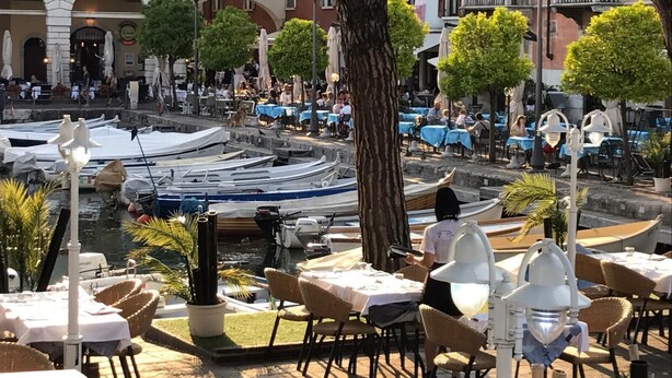 A riverside dining area with tables and chairs, boats docked along the water, and buildings in the background. - Atico Hotel (Desenzano del Garda)