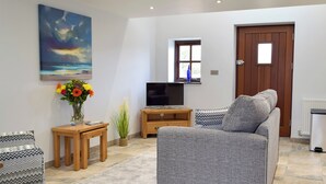 Una sala de estar con un sofá gris, una mesita de madera con un jarrón de flores, un televisor sobre un mueble de madera y un cuadro de un atardecer en la pared.