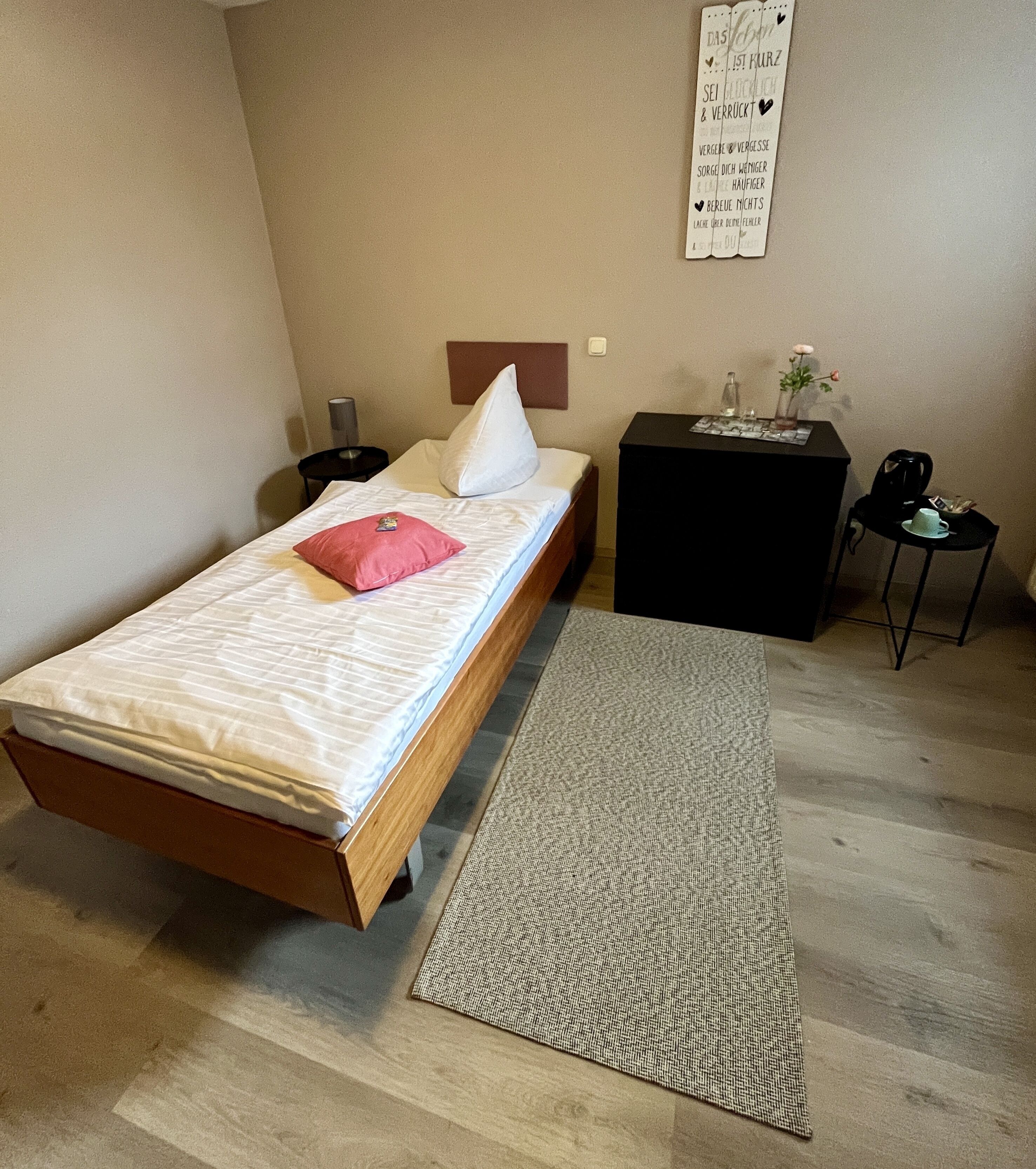 A bedroom with a bed, a bedside table, a nightstand, a small table with a vase, and a wall with text.