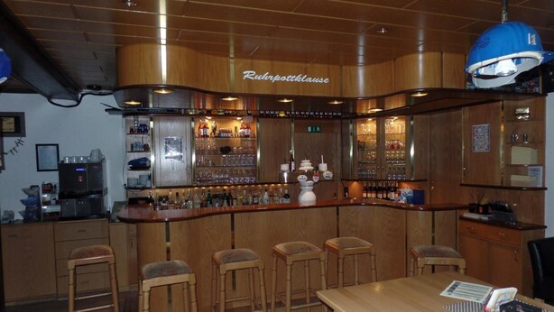 A bar area with wooden cabinets, bar stools, and a counter displaying various bottles and glasses. - Hotel Vogt (Bad Driburg)
