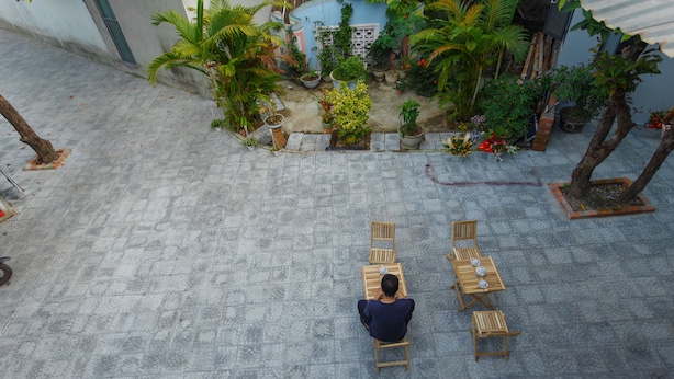 A courtyard with a person sitting on a chair, surrounded by potted plants and a few wooden tables. - King Homestay (Hue)