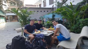 Trois personnes sont assises à une table à l’extérieur, devant des assiettes remplies de nourriture, entourées de verdure et de bâtiments.