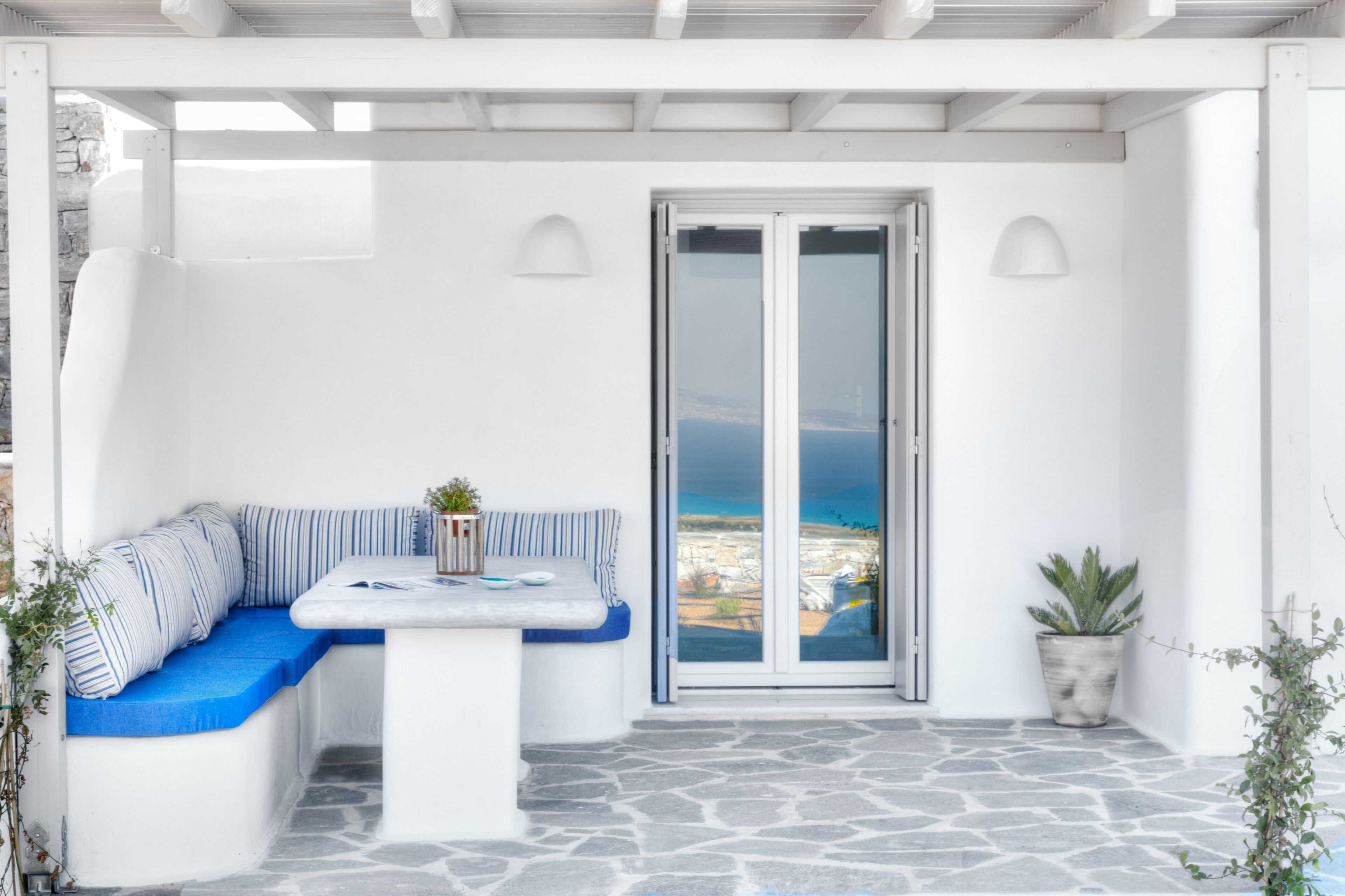 Un espacio exterior con asientos blancos, una mesa, un banco acolchado azul y una planta en maceta.