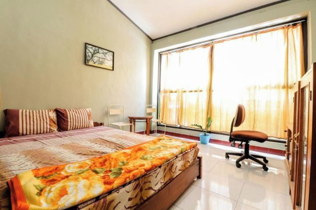 A bedroom with a large bed, a desk, a chair, and a window with curtains.