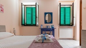 A hotel room with a bed, a desk, a chair, a mirror, and two windows with green shutters. - A Country House near the Lake Cod. 012104-CNI-00006 IT012104C24MJ5EPRC (Monvalle)