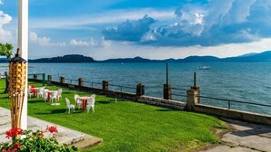 A waterfront terrace with tables and chairs, a grassy area, and a view of the water and distant mountains. - A Country House near the Lake Cod. 012104-CNI-00006 IT012104C24MJ5EPRC (Monvalle)
