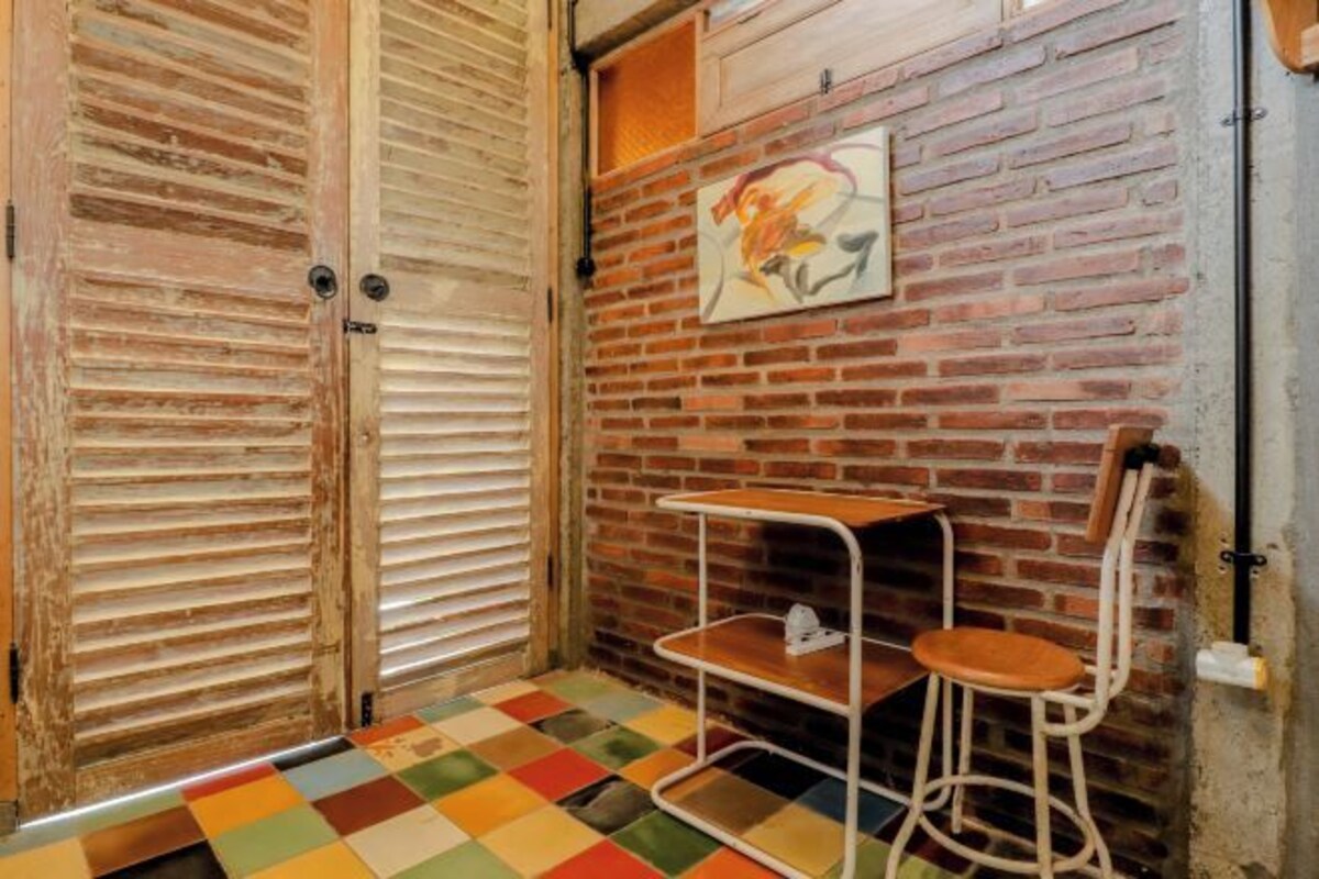 A room with a brick wall, a wooden door, a colorful checkered floor, a small white metal shelf, and a single wooden chair.