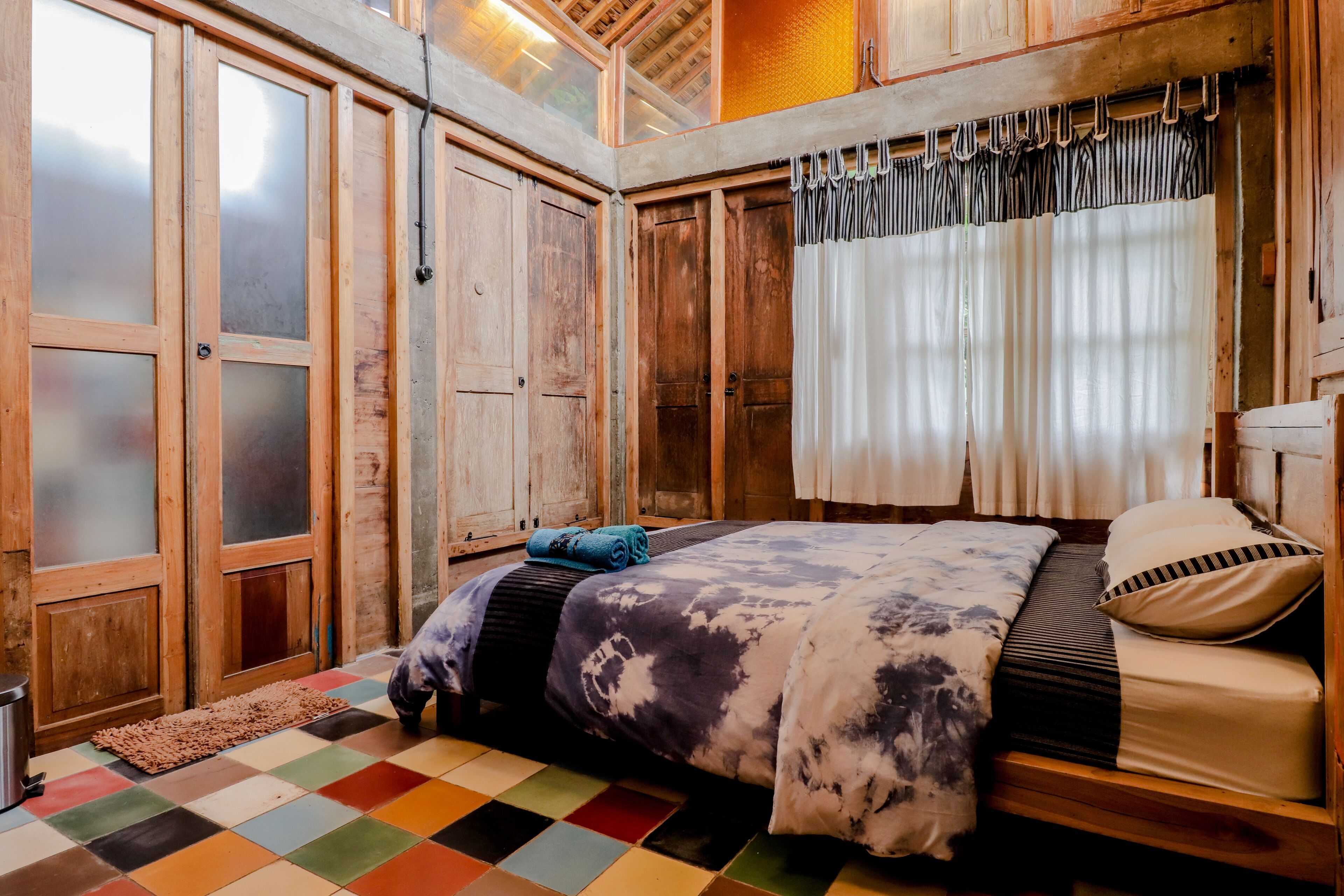 Une chambre avec un lit en bois, un sol à carreaux aux couleurs vives et une grande fenêtre avec des rideaux.