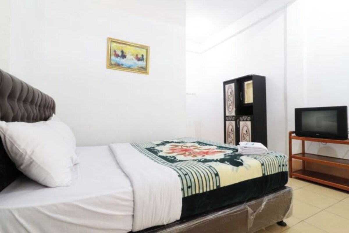 Una cama bien hecha con una colcha floral, cabecera negra y almohada blanca. A la derecha, un televisor sobre un mueble de madera.