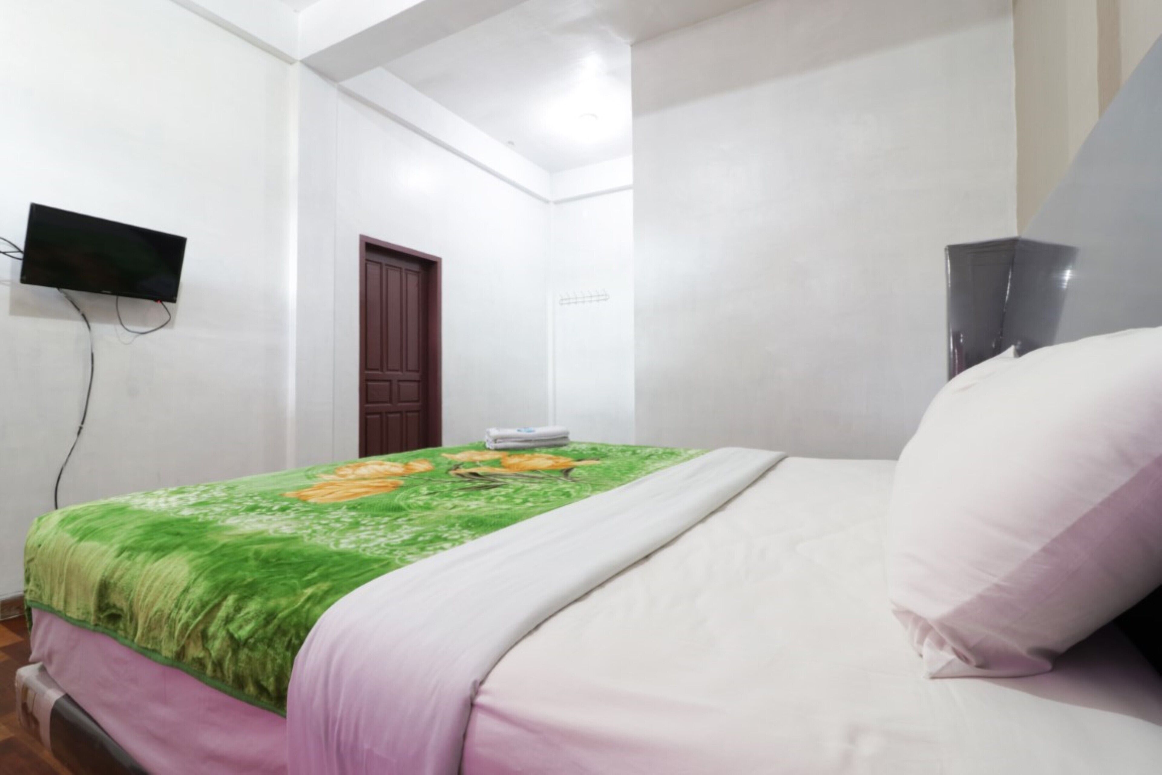 A bedroom with a bed, a flat-screen TV mounted on the wall, a door, and a small cabinet.