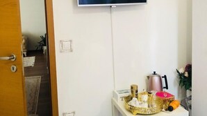 Ein Zimmer mit an der Wand befestigtem Fernseher, einem kleinen Tisch mit diversen Gegenständen, einer Tür und einer sichtbaren Treppe.