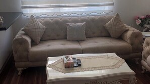 Ein Wohnzimmer mit einem beigen Sofa, einem runden Couchtisch und einem Teppich mit Muster, sowie einem Fenster mit Jalousien.