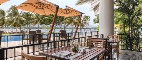 Una terraza con mesas y sillas, palmeras y vistas al mar.