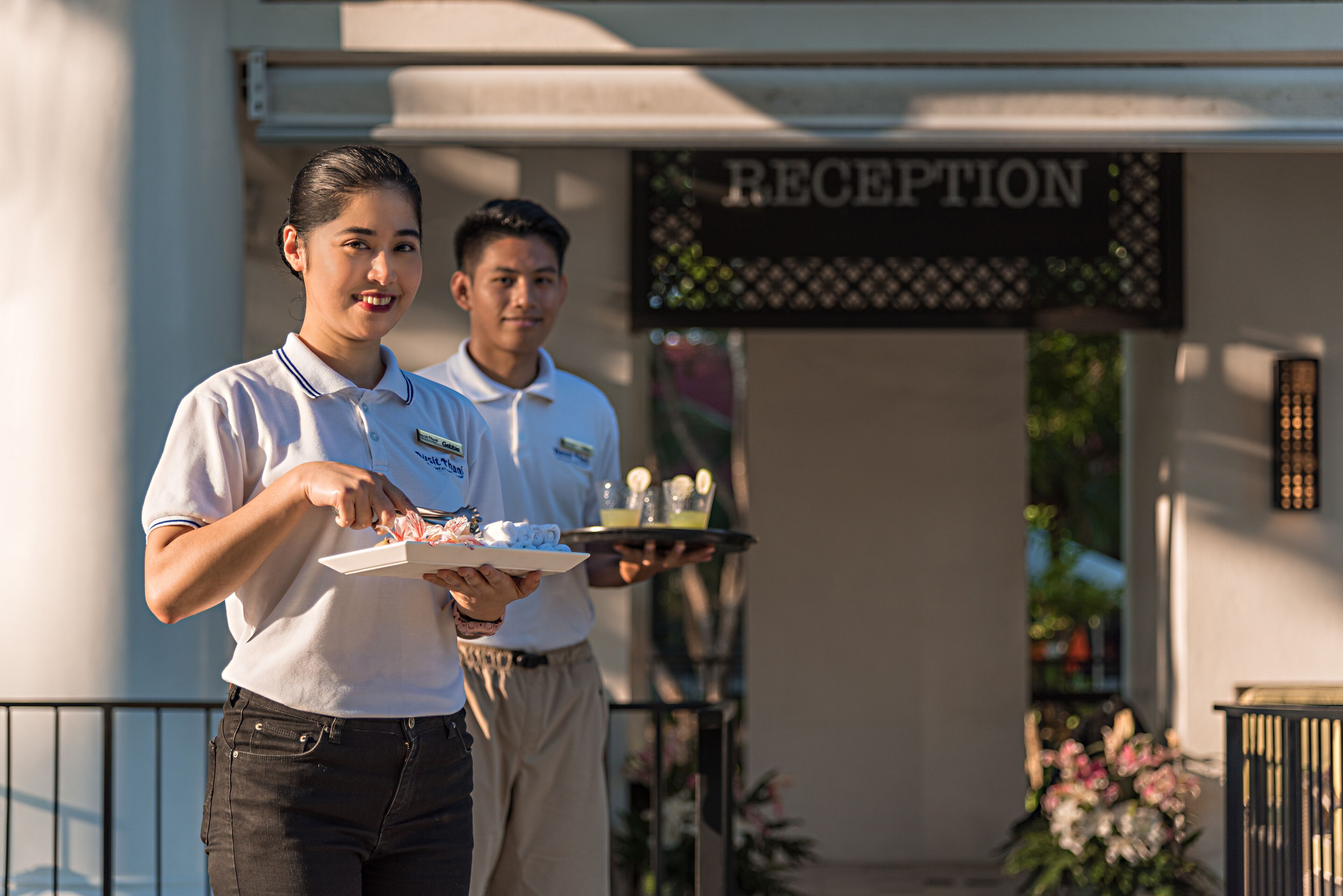 Two individuals in uniform holding trays with food and drinks, standing in front of a reception area.