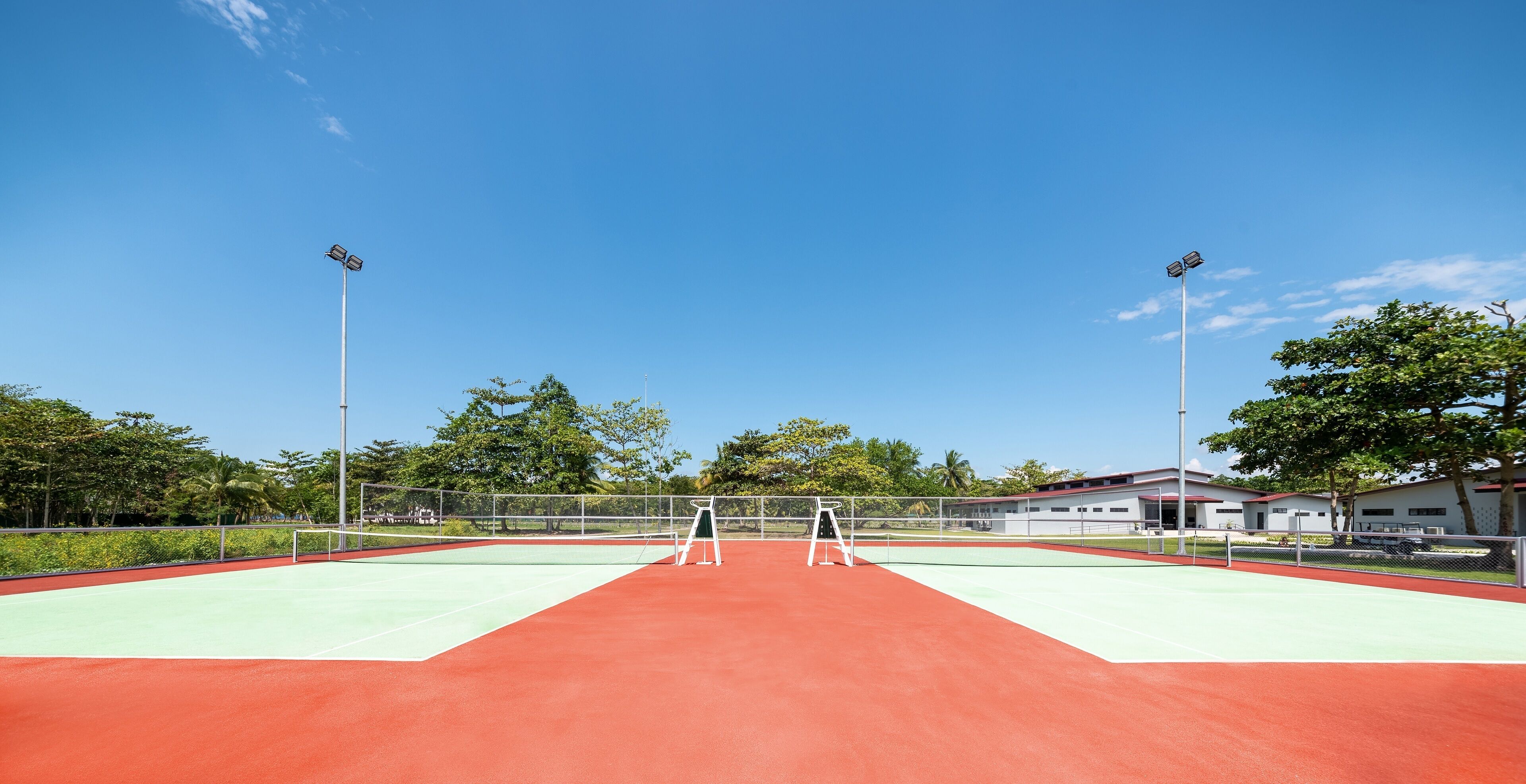 An outdoor tennis court with a red surface, surrounded by a fence, and two floodlights.
