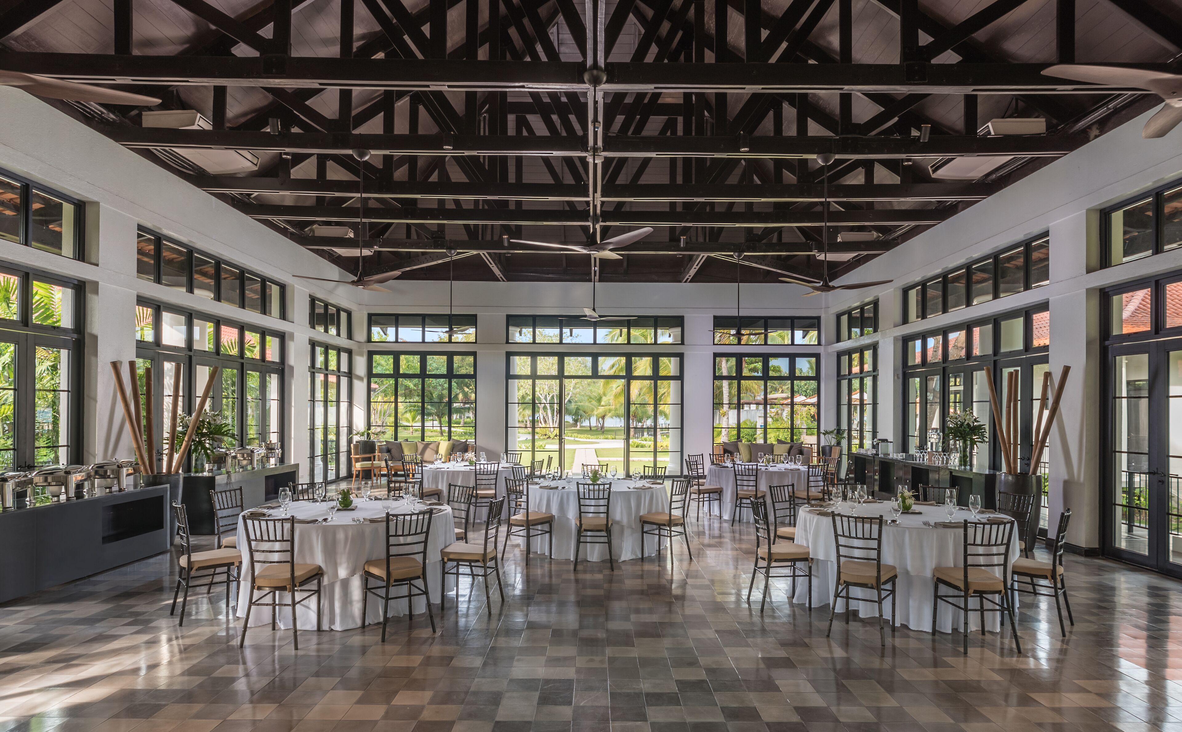 A spacious hall with multiple round tables and chairs, large windows offering views of a garden, and a high ceiling with exposed beams.