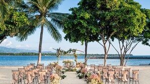 Une cérémonie de mariage organisée sur la plage, avec des chaises disposées en rangées, une arche décorée de fleurs et des palmiers.