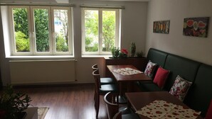 Een eetgedeelte met een houten tafel en stoelen, een groene bank en twee ramen met uitzicht op groen.