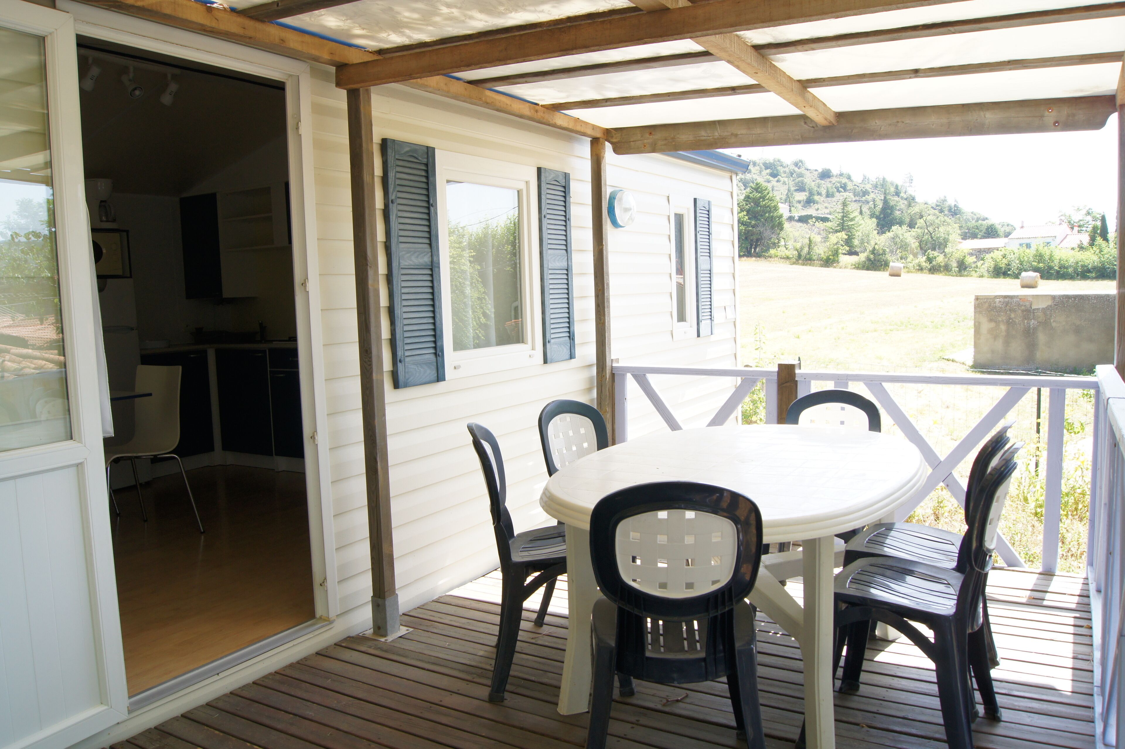 Un espace extérieur abrité avec une table blanche et des chaises noires, une terrasse en bois et un bâtiment blanc avec des volets bleus.