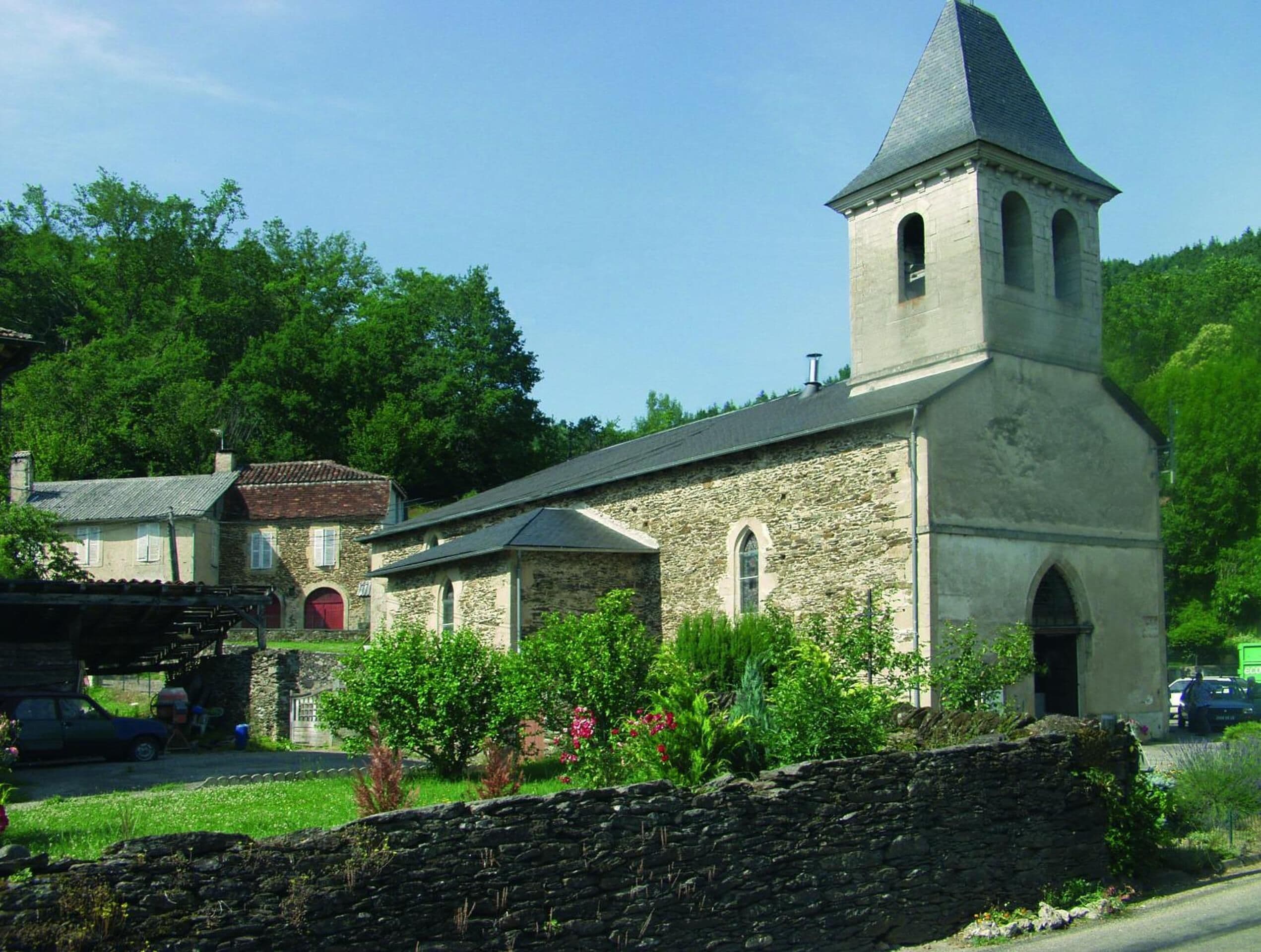 A stone church with a bell tower, surrounded by greenery and a stone wall.