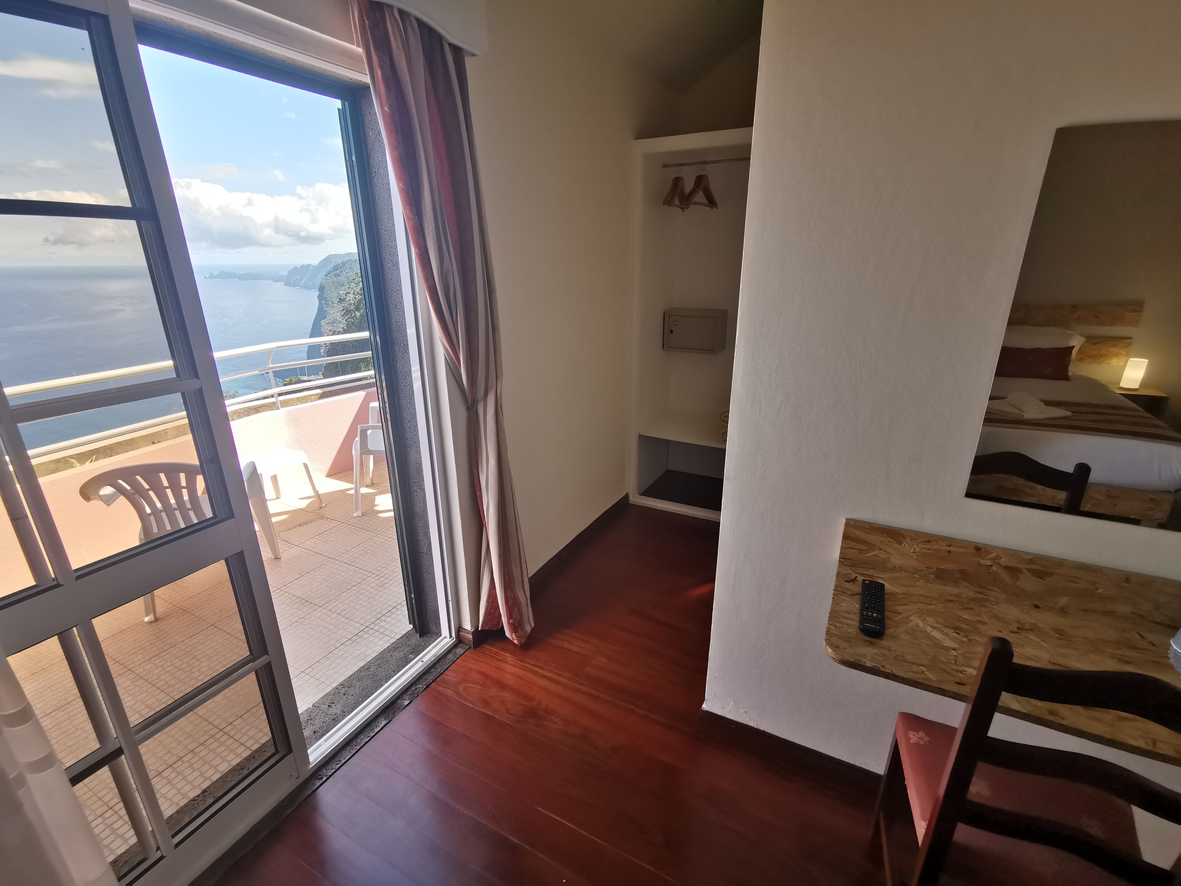 A room with a wooden floor, a bed with a wooden headboard, a small table with a remote control, and a view of the sea through a window with curtains.