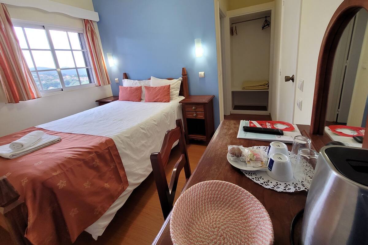 A hotel room with a bed, a desk with a tray of breakfast items, a wardrobe, and a view of the outdoors.