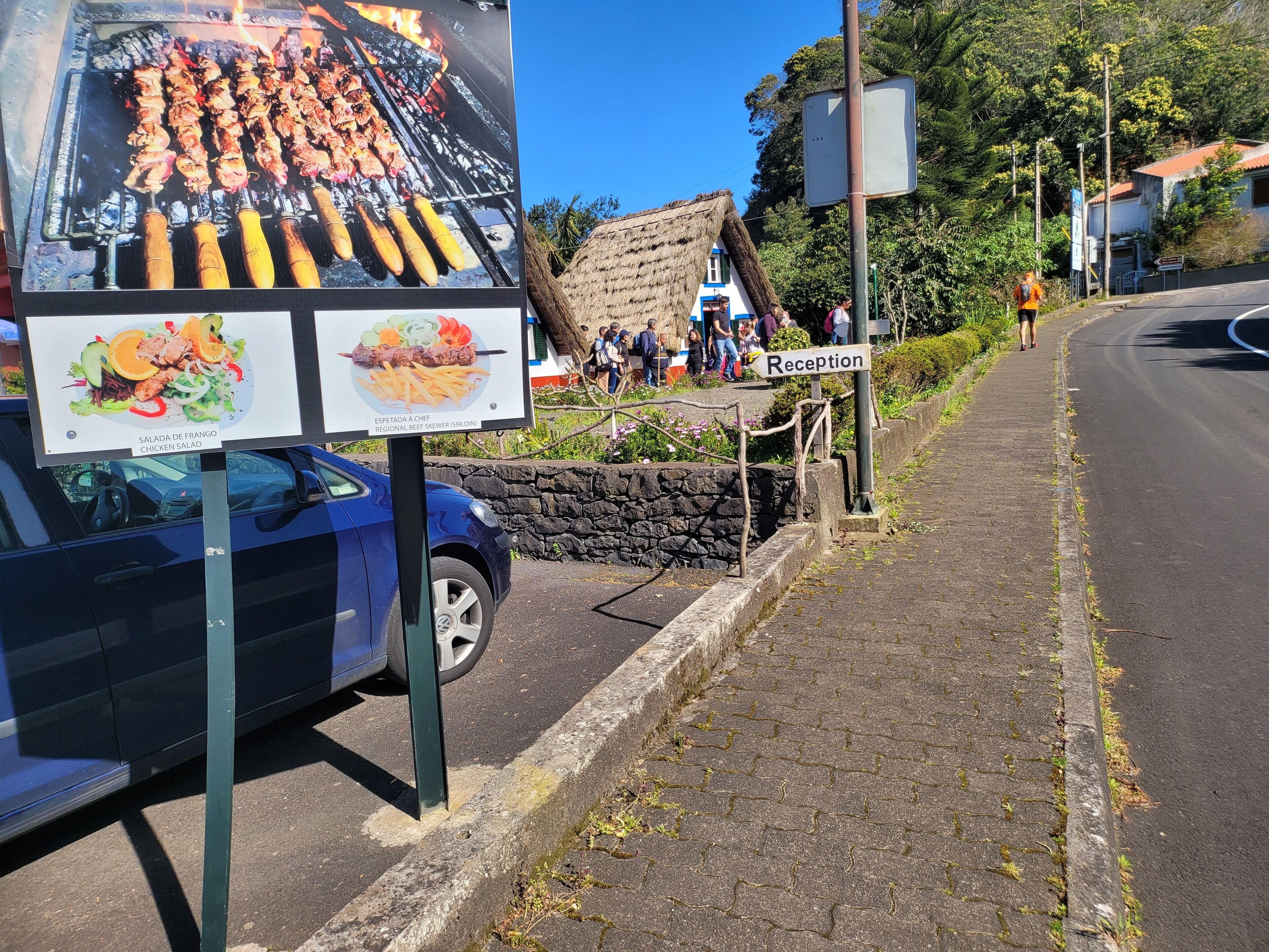 Un área de recepción con un letrero, un coche azul y un edificio con techo de paja.