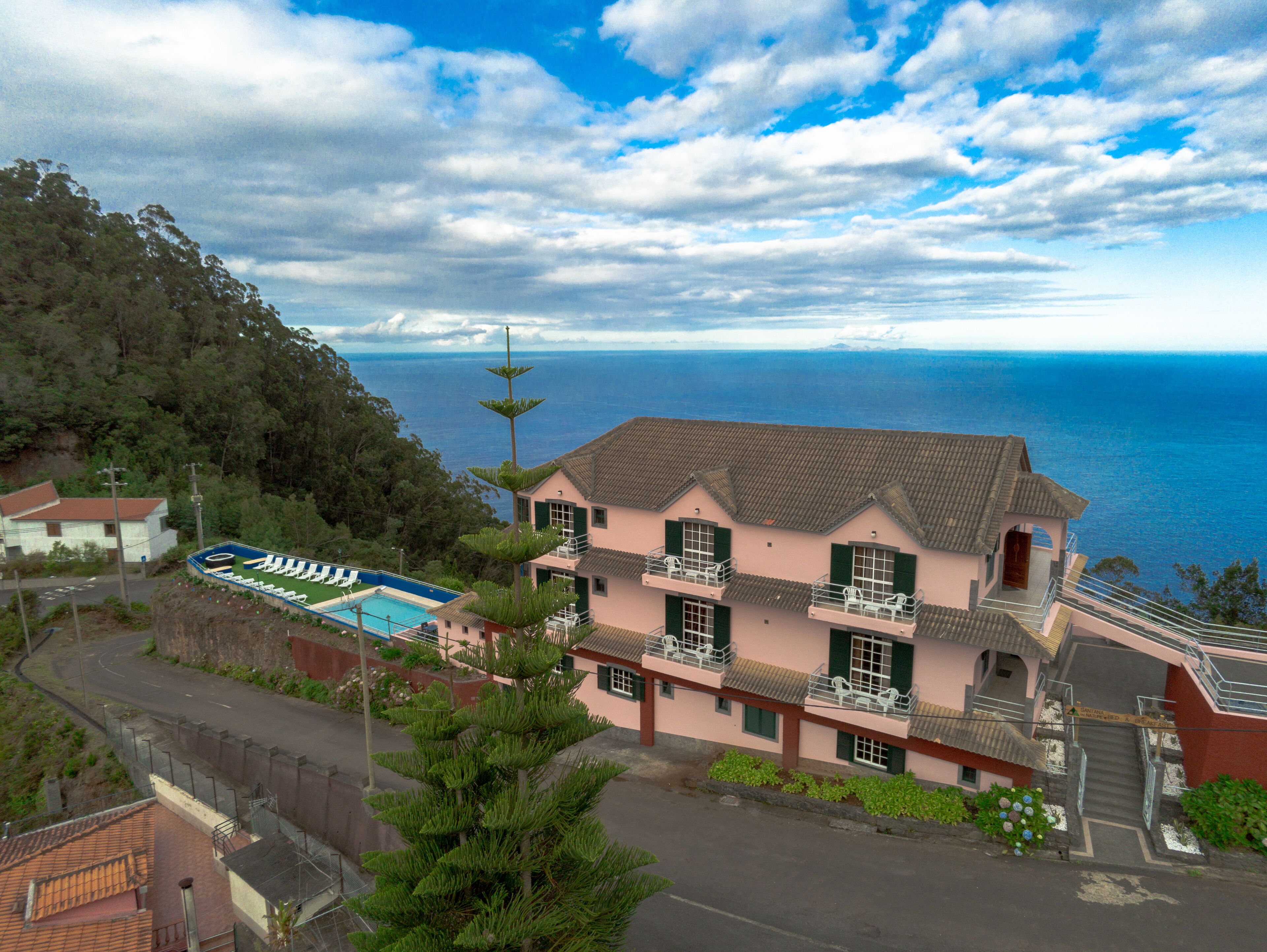 A two-story building with a swimming pool, a view of the sea, and a forested hillside.