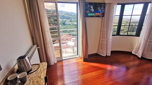 A room with a wooden floor, a TV mounted on the wall, a balcony with a view of the landscape, and a table with a kettle and cups. - Santana in Nature Bed & Breakfast (Santana)