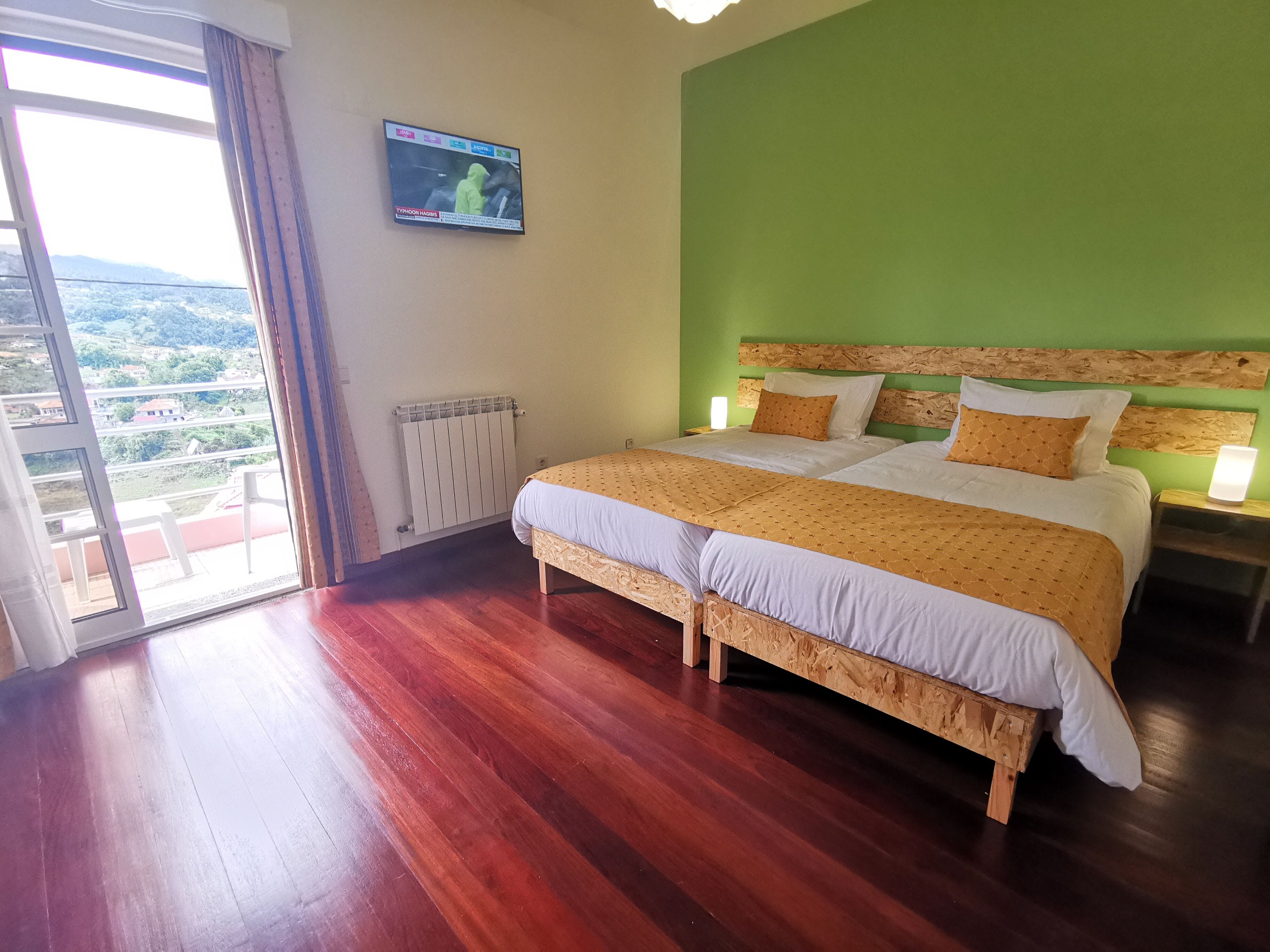 Un lit double avec une tête de lit en bois, deux tables de chevet avec des lampes, un téléviseur fixé au mur et une vue sur un paysage verdoyant par une grande fenêtre.