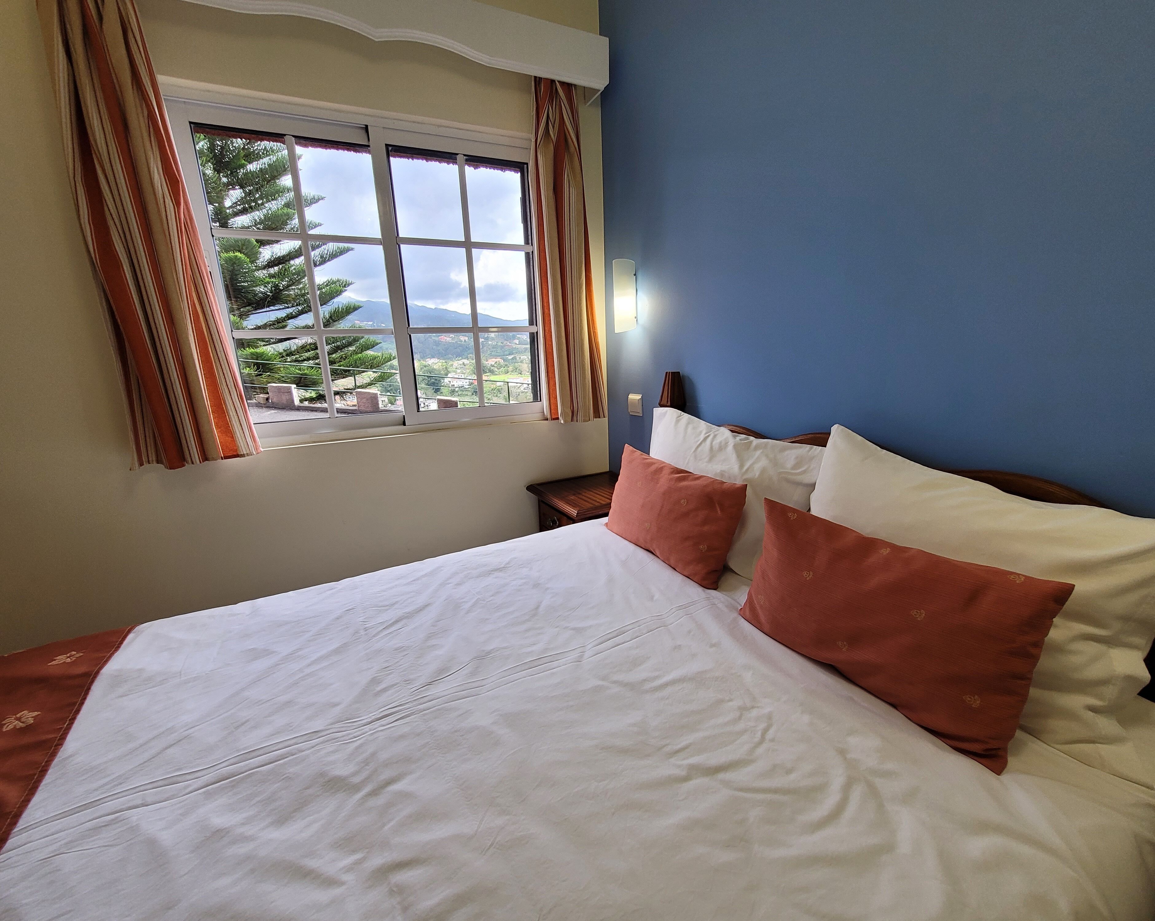 Una cama bien hecha con almohadas blancas y marrones, una ventana con cortinas y vistas al verde exterior.