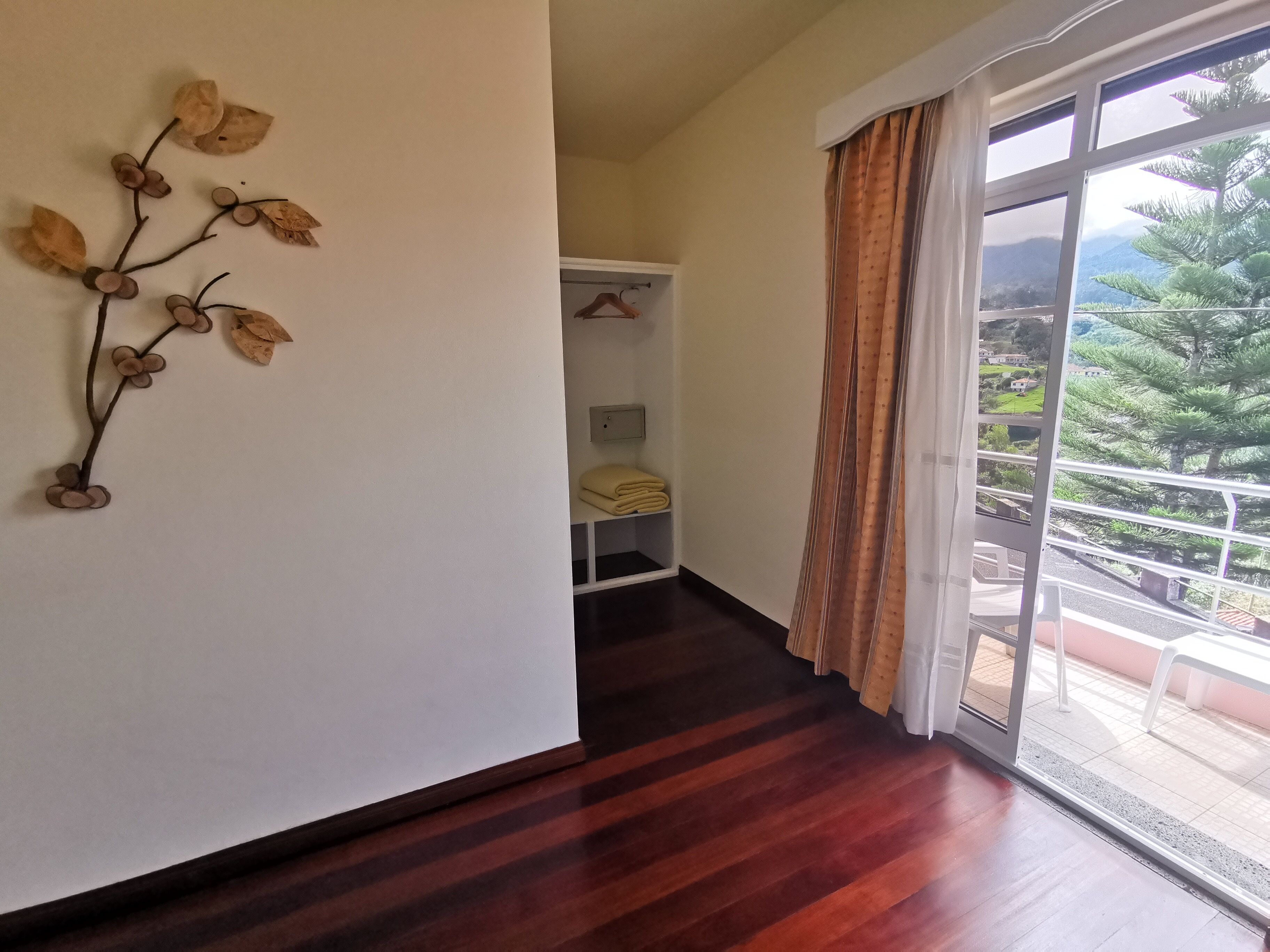 A room with a wooden floor, a large window with a view of greenery, and a wall-mounted decoration.