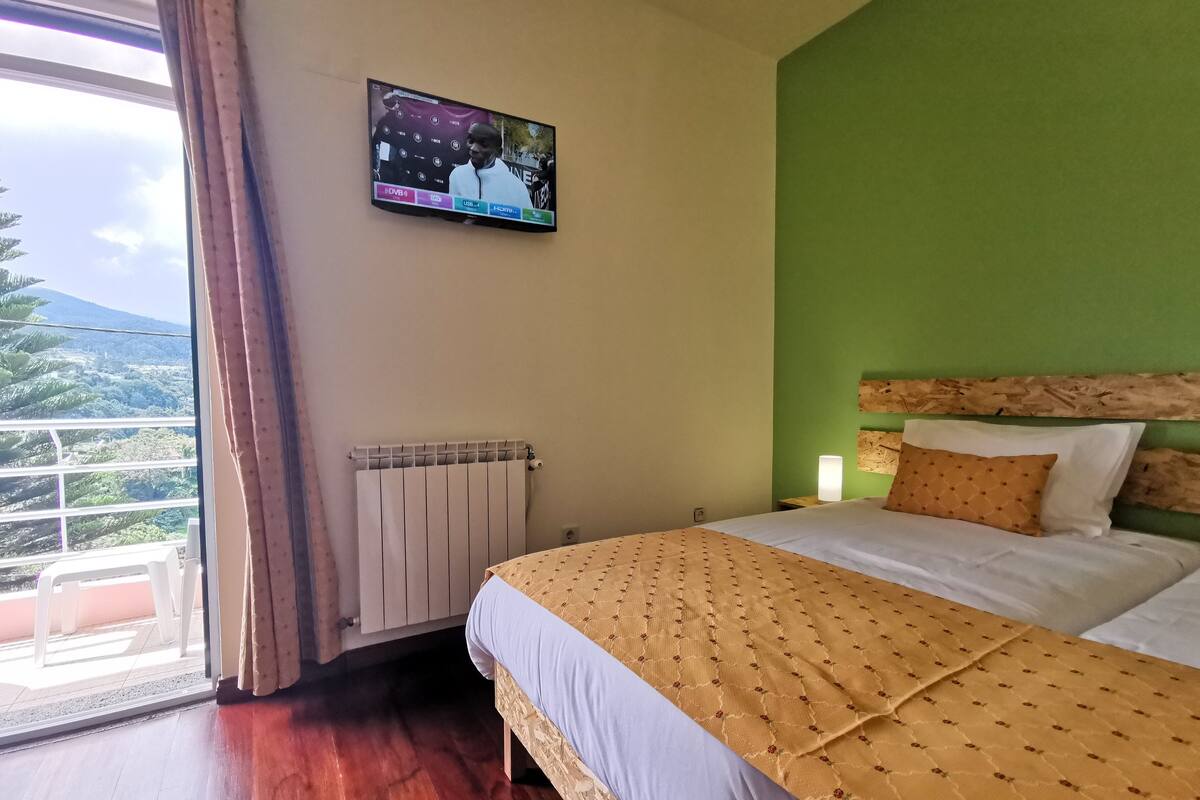 A hotel room with a bed, a television mounted on the wall, a radiator, and a window with curtains.