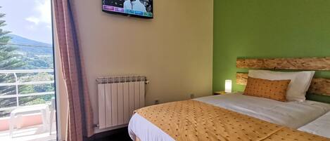 Una habitación de hotel con una cama, televisión montada en la pared, un radiador y una ventana con cortinas.