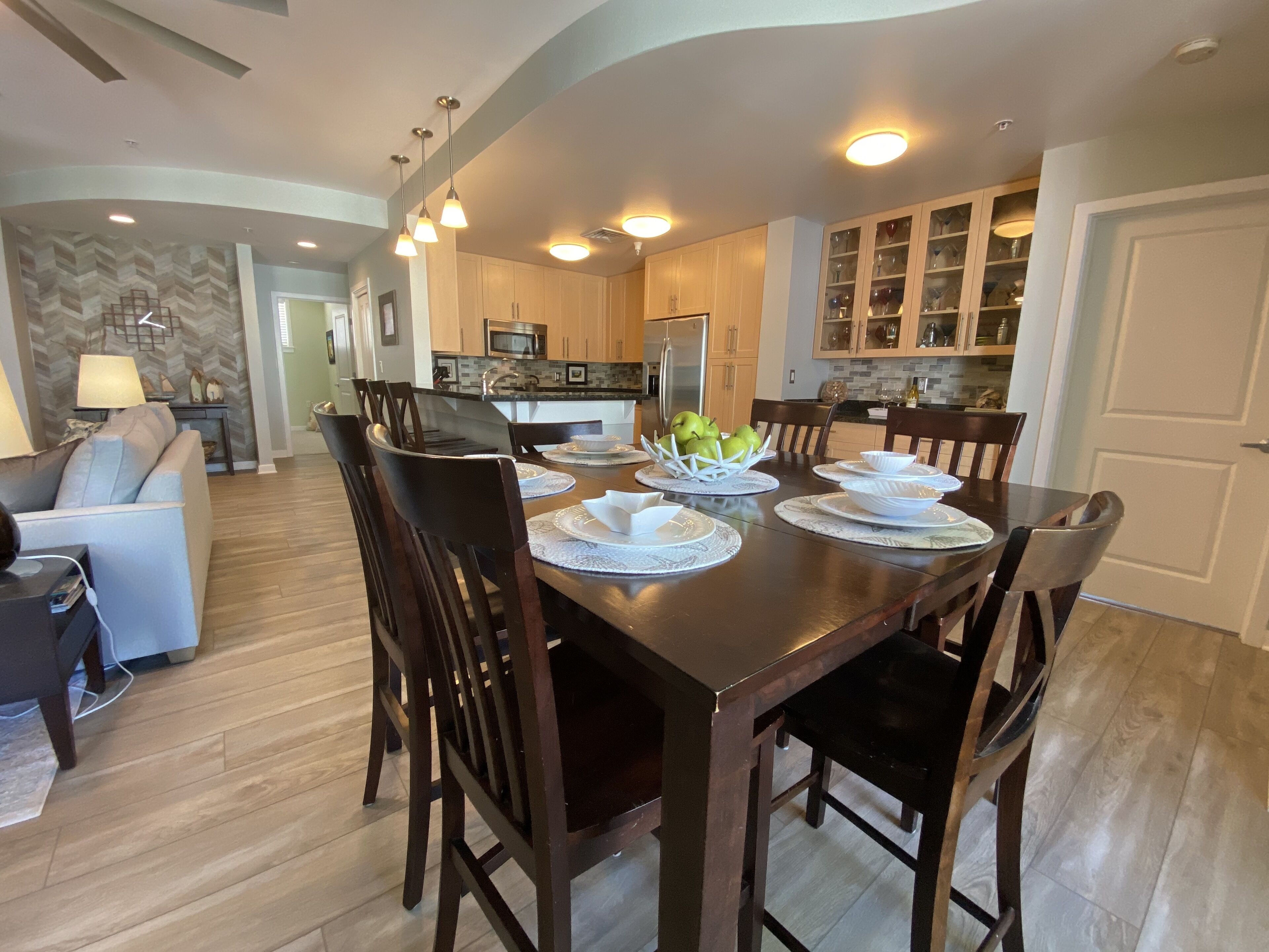 A dining area with a wooden table set for six, surrounded by chairs. There is a kitchen in the background with wooden cabinets and a microwave.