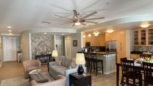 A spacious living room with a sofa, chairs, and a coffee table. There is a kitchen area with a refrigerator, microwave, and dining table with chairs. - Sandbridge Beach Condo with fantastic Ocean View and Bay View Balcony!  (Virginia Beach)
